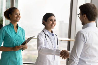 Çok ırklı genç doktor kadınlar tıbbi patronla el sıkışıyor, gülümsüyor, gülüyor. Pozitif çeşitlilikte hastane meslektaşları el sıkışıyor, başarılı bir işbirliği tartışıyorlar.