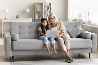Akıllı büyükannenin çocuklara uygulamayı dizüstü bilgisayarda kullanmayı öğretmesi, okul ödevlerine yardım etmesi, modern iç mekanlarda bilgisayar başında rahat bir koltukta oturması.