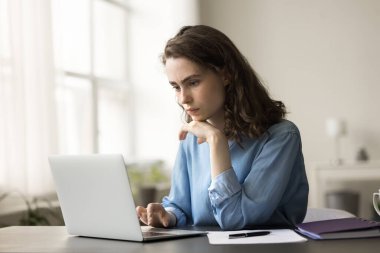 Odaklanmış genç serbest çalışan profesyonel kız evde online proje üzerinde çalışıyor, dizüstü bilgisayarla masada oturuyor, çeneye dokunuyor, daktilo ediyor, internet teknolojisi kullanıyor.
