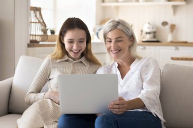 Mutlu genç kız ve neşeli büyükanne evde kanepede eğleniyor, dizüstü bilgisayarı tutuyor, film izliyor, online konferansta konuşuyor aile görüşmesi, gülümsüyor, gülüyor, eğleniyor, iletişim kuruyor.