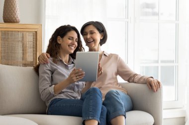 Gülen anne ve 35 'lerin kızı yeni uygulamanın keyfini çıkarıyorlar, modern dijital tablet cihazını kullanarak oturma odasında kanepede oturuyorlar, video izliyorlar, çok nesilli aile internette eğleniyor.