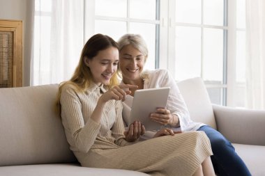 Olumlu akıllı genç kız ve oldukça olgun büyükanne evde modern dijital teknolojinin tadını çıkarıyorlar, online servisi kullanıyorlar, internet iletişimi için tablete başvuruyorlar, gülümsüyorlar, gülüyorlar.