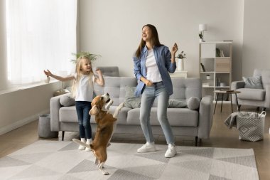 Heyecanlı bir anne ve neşeli bir evcil hayvanla dans eden mutlu bir anaokulu çocuğu, oturma odasında hoplayıp zıplıyor, eğleniyor, eğleniyor, eğleniyor, evde müzik dinliyor, sevimli bir av köpeği eğitiyor. Tam uzunluk