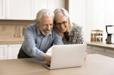 Mutlu yaşlı koca ve eş ev mutfağındaki dizüstü bilgisayarda online uygulama kullanıyor, daktilo çekiyor, ekrana bakıyor, gülüyor, emekliliğin tadını çıkarıyor, internet iletişimi kuruyor.