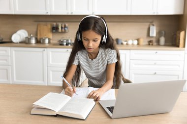 Kablosuz kulaklıklarla evde ders çalışan, dizüstü bilgisayar kullanan, video eğitimi alan, defter defterine not yazan, mutfak masasında duran liseli kız çocuğuna odaklan.