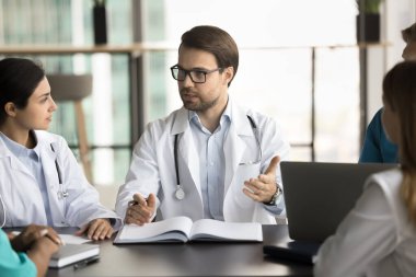 Gözlüklü ve beyaz önlüklü ciddi bir erkek doktor çeşitli personelle yaptığı toplantıda konuşuyor, çok ırklı profesyonel meslektaşlarıyla konuşuyor, tıp seminerlerinde sağlık hizmetleri işini tartışıyor.