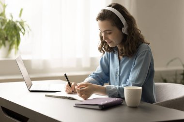 Modern nesil Z genç yetişkin kız kulaklıklarıyla bilgisayarda çalışıyor, ses konferanslarını dinliyor, web sitesi kuruyor, notlar yazıyor, ofis masasında oturuyor.