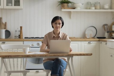 Evde çalışan, dizüstü bilgisayarda yazan, mutfak masasında oturan ciddi bir iş kadını. Ev sahibi kadın internet uygulaması kullanarak ev ödemesi yapıyor.