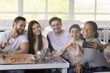 Kafede akıllı telefondan selfie çeken, kendini videoya çeken, cep telefonu tutan, gülümseyen, gülen, pizzayla masaya poz veren, sıcak içecekle eğlenen mutlu arkadaşlar.