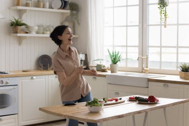 Neşeli, kıdemli şef kadın mutfak masasında dans edip şarkı söylüyor, taze sebzelerle salata pişiriyor, sağlıklı ve aktif bir yaşam tarzı sürdürüyor, evde eğleniyor, aşçılık hobilerinden keyif alıyor.
