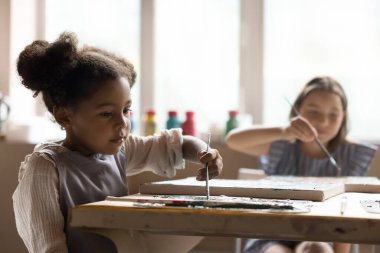 Önlüklü güzel kız öğrenci yaratıcı atölye dersleri alıyor, masadaki tuvalde resim yapıyor, boya fırçası ve palet kullanıyor, sanatsal yetenekleri eğitiyor, çizim becerileri geliştiriyor.