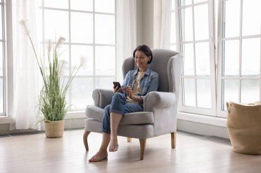 Evde cep telefonuyla internet uygulaması kullanan, rahat, geniş pencereli şık bir evde rahat bir koltukta dinlenen, daktilo kullanan, internette sohbet eden olgun bir kadın.
