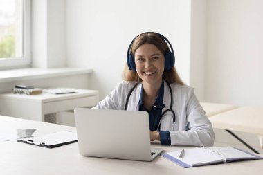 Ofisteki dizüstü bilgisayarında oturan, kameraya bakan, gülümseyen, uzaktan kumandalı online danışmayı destekleyen, hastalarla internet iletişimini sağlayan kulaklıklı mutlu doktor kadın. Kadın portresi