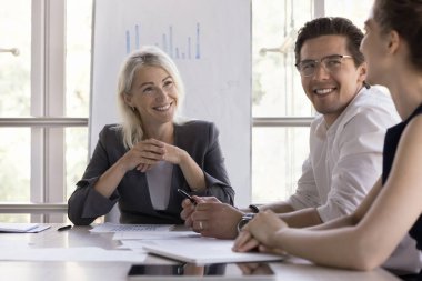 Genç çalışanların fikirlerini dinleyen mutlu orta yaşlı kadın lider beyin fırtınası toplantısı öneriyor, gülümsüyor, gülüyor. Küçük işletme ekibi masada projeyi tartışıyor. Akıl hocası stajyer.