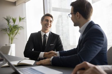 İki mutlu iş adamı el sıkışıyor, konferans masasında oturuyor, kapanış toplantısı, sözleşme, başarılı proje, konuşuyor, gülüyor, iş görüşmesinden sonra tokalaşıyor.