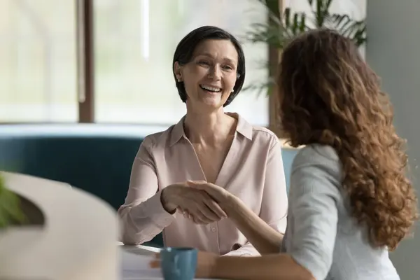 Positive Senior Business Woman Giving Handshake Younger Colleague Work Table Royaltyfrie stock-billeder
