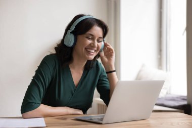 Büyük parlak kulaklıklı mutlu serbest çalışan kadın video görüşmeleri yapıyor, dizüstü bilgisayarda konuşuyor, gülümsüyor, gülüyor, çevrimiçi iletişimden keyif alıyor. Müzik dinleyen serbest çalışan.