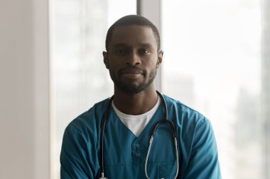 Üniformalı üniformalı, klinikte poz veren, kameraya bakan kendinden emin, genç Afrikalı cerrah. Pozitif ciddi siyah erkek doktor, pratisyen portresi