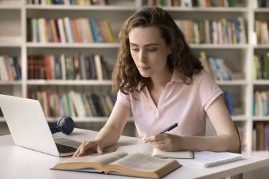 Ciddi bir şekilde kampüs kütüphanesinde okuyan, kitap okuyan, masada ders kitabı yazan, makale, makale, dizüstü bilgisayarla tdesk 'te araştırma yapan güzel bir kız.