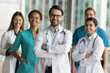 Steteskoplu mutlu doktor adam arka planda yan yana duran çeşitli tıbbi meslektaşlarıyla birlikte grup fotoğrafı için poz veriyor, elleri çaprazlanmış, gülümseyerek kameraya bakıyor.