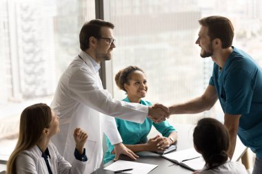 Zor tıbbi hasta davasında başarılı işbirliğini kutlayan iki mutlu doktor, masanın üstünde el sıkışıyor, gülümsüyor, gülüyor, çok ırklı çalışma arkadaşlarıyla buluşuyor.