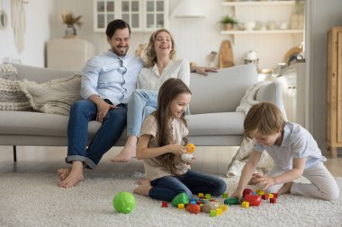 Oyuncak bloklarla oynayan iki neşeli, heyecanlı kardeş çocuk, yumuşak halı döşeli zeminde renkli küpler, kuleler inşa ediyorlar, plastik parçaların üzerinde gülüyorlar. Bir çift ebeveyn küçük çocuklara bakıyor.