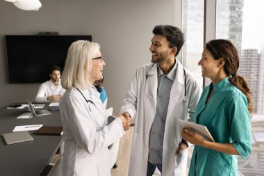 Beyaz önlüklü neşeli kıdemli doktor kadın çeşitli genç meslektaşlarıyla el sıkışıyor başarılı stajyerler, kahkahalar atıyor, hastane yönetim kurulundaki tıp konferansında iletişim kuruyor.