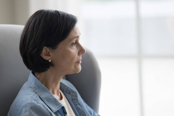 Sad pensive mature black haired woman looking away in deep thoughts, thinking of future retirement, loss, emotional crisis, health problems, sitting on couch, armchair at home