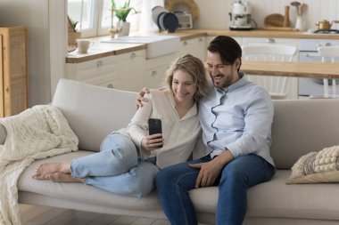 Neşeli, genç bir çift kanepesinde sarılıyor, cep telefonu tutuyor, internet üzerinden iletişim için başvuru yapıyor, video görüşmesinde konuşuyor, cihaz ekranına bakıyor, gülümsüyor, gülüyor.