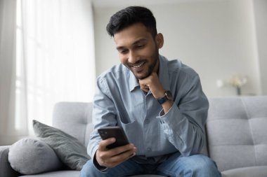 Neşeli, genç, yetişkin Hintli adam cep telefonu tutuyor, aletlerle eğleniyor, eğleniyor, sohbet ediyor, çevrimiçi video görüşmeleri yapıyor, okuyor, internet içeriğini izliyor.