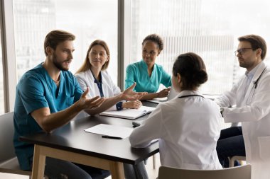 Mavi sıhhiye üniformalı kendinden emin genç cerrah hastane toplantı odasında çok ırklı meslektaşlarıyla konuşuyor, masadaki çeşitli doktorlarla konuşuyor, klinik ekip çalışması için fikirler sunuyor.