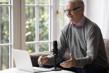 Mikrofon ve dizüstü bilgisayarda konuşan, video görüşmesinde konuşan, gülümseyen, gülen yaşlı bir adam. Kıdemli öğretmen, nüfuz sahibi Webinar, evden online ders veriyor.