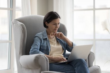 İş iletişimi için online uygulama kullanan, evde çalışan, ekrana bakan, okuyan, izleyen, düşünceli bir gülümsemeyle çeneye dokunan olgun, serbest çalışan bir kadın.