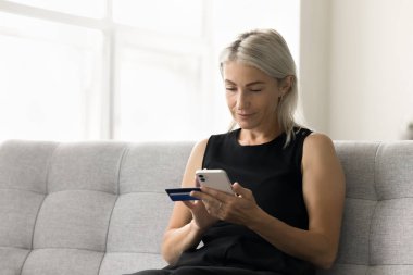 Odaklanmış olgun banka müşterisi kadın cep telefonunda bankacılık uygulaması kullanıyor, elinde mavi kredi kartı tutuyor, internet mağazalarından alışveriş yapıyor, online ödeme yapıyor.