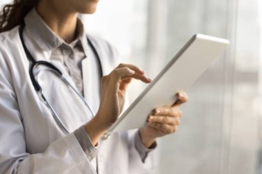 Beyaz üniformalı genç bayan doktor dijital tablet tutuyor, online danışmanlık yapıyor, modern teknolojiyi kullanıyor, iş için internet iletişimi kuruyor, elektronik kayıt yapıyor. Kırpılmış atış