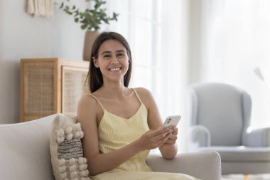 Neşeli, genç, akıllı telefon kullanıcısı kız cep telefonu tutuyor, kameraya bakıyor, gülümsüyor, gülüyor, kanepede dinleniyor, çevrimiçi iletişimin tadını çıkarıyor, kablosuz bağlantı teknolojisi kullanıyor. Ev resmi