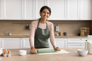 Önlüklü güzel fırıncı kadın ev portresi. Neşeli genç bir kadın mutfakta pasta pişiriyor, dişlek gülüşüyle kameraya bakıyor, ev işlerinden hoşlanıyor, tatlı hazırlıyor.