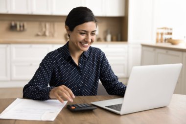 Mutlu Hintli serbest muhasebeci kadın evde muhasebe yapıyor, kar sayıyor, yatırım gelirlerini hesaplıyor, online finans uygulamasını kullanıyor, internet teknolojisini dizüstü bilgisayarda kullanıyor, gülümsüyor, gülüyor.
