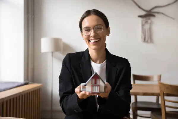 stock image Happy young real estate agent offering property buying and selling service, legal consultation about mortgage, insurance, help with rent, showing toy house at camera, laughing, posing for portrait