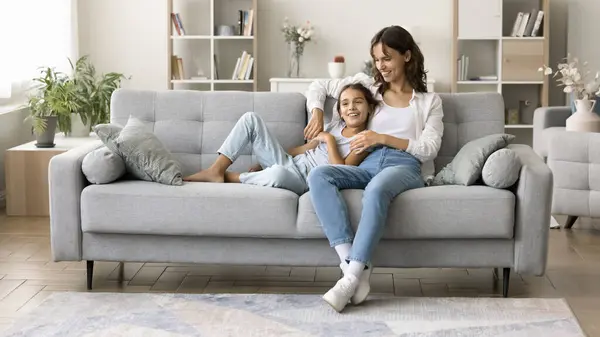 stock image Happy young mom and sweet kid girl enjoying homey leisure on comfortable couch, talking, smiling, looking away, resting on sofa together in modern living room, stylish home interior. Banner shot