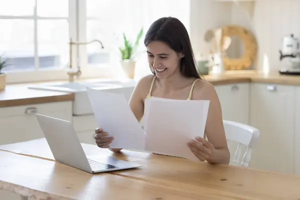 stock image Happy young Latin woman reading paper document at home, doing paperwork at table with laptop computer, smiling, laughing, getting letter with good news, celebrating success