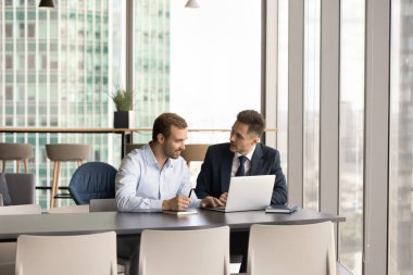 İki pozitif iş ortağı çağdaş şehir ofisindeki iş yerinde büyük pencereli, işbirliği, ortaklık, dizüstü bilgisayardaki online proje hakkında konuşuyor. Müdür danışman iş arkadaşı