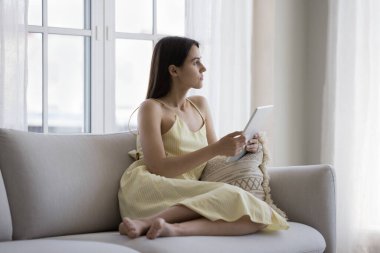 Dijital cihazlar taşıyan, düşünceli, düşünceli, genç bir kız öğrenci kanepeye oturmuş derin düşüncelere dalıp, düşünüyor, karar veriyor, keyfine bakıyor, internet iletişiminden keyif alıyor.