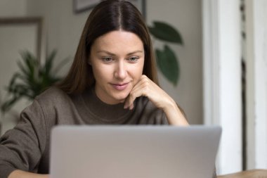 Ofiste dizüstü bilgisayarda oturan düşünceli, serbest çalışan bir kadın, çenesini tutarak online proje sunumunu izliyor, internet içeriğini okuyor, müşterilerle iletişim kuruyor.