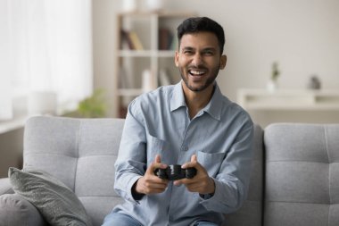Evde video oyunu oynayan neşeli genç Hintli oyuncu, elinde oyun sahası, joystick, oyun kumandası, gülüyor, bağırıyor, kanepeye oturuyor, kameraya bakıyor, boş zamanların tadını çıkarıyor, sanal aktivitelerin tadını çıkarıyor.