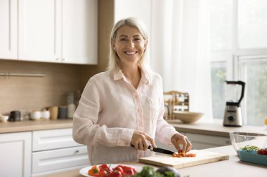 Ev mutfağında güzel şef kadın portresi. Pozitif sarışın bayan akşam yemeği, öğle yemeği için ev yemeği hazırlıyor, taze sebzeleri doğruyor, kameraya bakıyor.