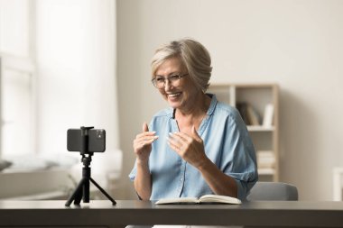 Neşeli kıdemli yaşlı nüfuzlu kadın blog için çekim yapıyor, evde selfie videosu kaydediyor, cep telefonu kamerasında konuşuyor, tripoda sabitlenmiş, gülümsüyor, çevrimiçi yayın yapıyor.