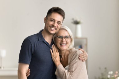 Olumlu yakışıklı oğul adam ve mutlu sarışın olgun anne evde sevgiyle sarılıyor, dişlek tebessümle kameraya bakıyor, gülüyor, aile fotoğrafı için yakın duruyor.