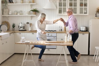 Neşeli yaşlı gri saçlı çift mutfak masasında dans ediyor, eğleniyor, gülüyor, şarkı söylüyor, müzikten hoşlanıyor, yemek pişiriyor, aile eğlencesi, aile aktivitesi, el ele tutuşuyor. Tam boy çekim