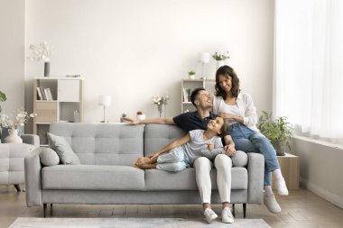 Mutlu bir çift ebeveyn ve küçük kız çocuğu, modern evdeki rahat koltukta oturup dinleniyor, konuşuyor, gülüyor, gülümsüyor. Tam boy geniş çekim